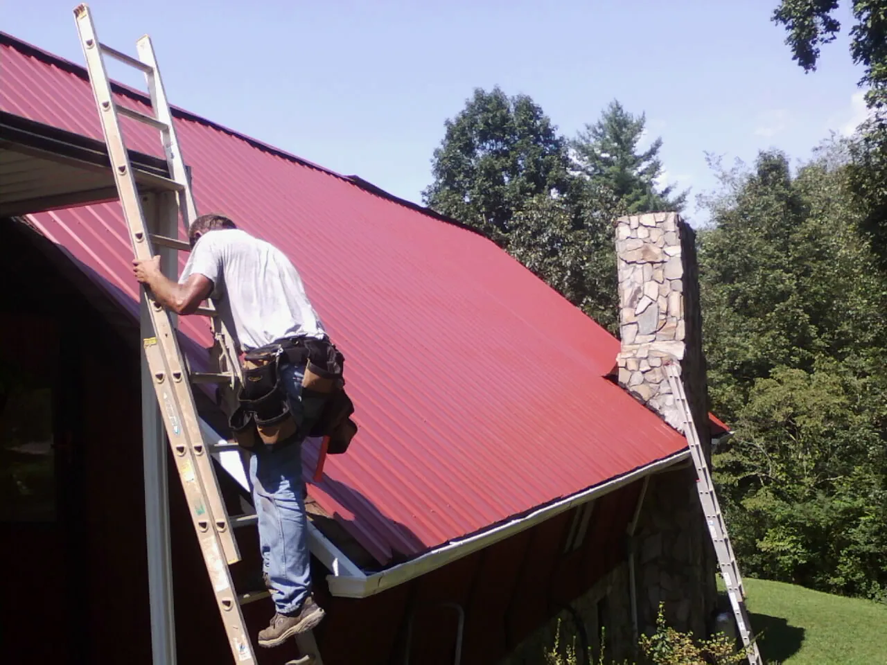 Roofer on a ladder beside a red metal roof and stone chimney — Soffit & Fascia Repair by Roofing Brothers in Brandy Station